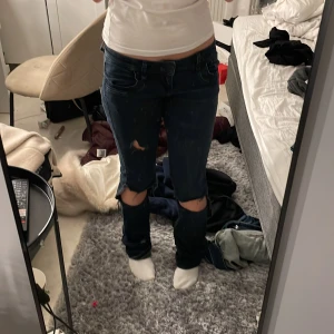 Mörkblå ripped jeans med låg midja - Säljer ett par mörkblå jeans med låg midja och slitna detaljer vid knäna. Byxorna har klassisk femficksdesign och är tillverkade i jeansmaterial. Passformen är rak och de har coola hål vid båda knäna för en edgy look.