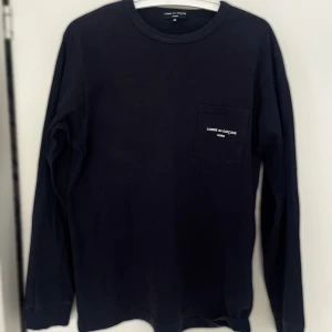 Mörkblå långärmad tröja Comme des Garçons Homme - Mörkblå långärmad tröja från Comme des Garçons Homme i storlek M. Tröjan har en rund hals och en bröstficka med vit broderad logotyp. Tillverkad i mjuk bomull, perfekt för en clean och stilren look.