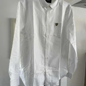 Vit skjorta från Lyle & Scott, storlek L NY - Vit skjorta från Lyle & Scott med klassisk krage och knappstängning hela vägen. På bröstet finns en ficka med den ikoniska broderade gula örnen. Skjortan är långärmad och tillverkad i mjuk bomull, perfekt för en clean och stilren look.