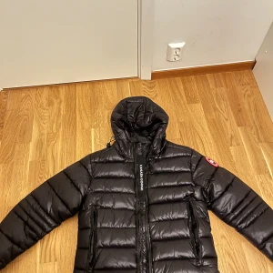 Svart pufferjacka från Canada Goose - Svart pufferjacka från Canada Goose med huva och klassisk logga på ärmen. Jackan har horisontella quiltade sektioner, två dragkedjeförsedda fickor och tydlig Canada Goose-tryck på dragkedjan. Perfekt för kalla dagar och har en snygg, modern look.