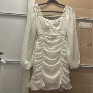 Vit klänning  - Säljer en elegant vit klänning från NLY Trend med långa ärmar och rynkade detaljer. Klänningen har en dragkedja i ryggen och en volangkant nedtill. Perfekt för stunden.