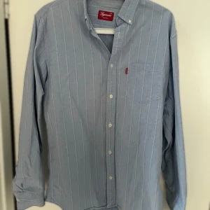 Blå randig skjorta från Supreme - Supreme oxford skjorta i ljusblått med vita vertikala ränder, klassisk krage och bröstficka med Supreme-tag. Skjortan har knappar hela vägen och är tillverkad i 100% bomull. Perfekt för dig som gillar streetwear med clean look. Finns en litet skada på kragen som syns på bilden. Skjortan är väldigt sällsynt gjort i Japan.
