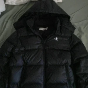 Svart pufferjacka från Calvin Klein Jeans - Svart pufferjacka från Calvin Klein Jeans med huva och dragkedja framtill. Jackan har ett stilrent CK-logo broderat på bröstet och är fodrad för extra värme. Perfekt för kalla dagar och har en modern, loose passform.