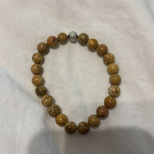 Brunt pärlarmband med silverdetalj - Coolt armband med runda bruna pärlor i natursten och en glittrig silverfärgad pärla som accent. Elastiskt band gör det enkelt att ta på och av. Perfekt accessoar för dig som gillar en jordnära och unik stil.