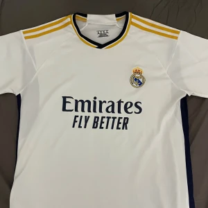 Real Madrid T-shirt stl  XL - Säljer en Real Madrid fotbollströja Adidas i vit polyester med gula ränder på axlarna och mörkblå detaljer. Tryck med klubbmärke och Emirates Fly Better på bröstet. Rund hals med gul och mörkblå kant. Perfekt för dig som älskar fotboll och Real Madrid.