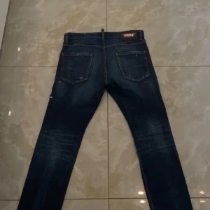 Mörkblå DSQUARED2 jeans med slitningar - Säljer ett par mörkblå jeans från DSQUARED2 med coola slitningar och detaljer på benen. Klassisk femficksmodell med smal passform och snygga kontrastsömmar. Perfekt för dig som gillar en edgy streetstil och vill sticka ut.
