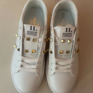 Valentino Rockstud vita sneakers - Säljer ett par vita Valentino Rockstud Untitled sneakers med coola guldfärgade nitar på sidorna och ovansidan. Skorna har snörning, rund tå och platt sula. Tillverkade i lyxigt skinn med klassisk Valentino-logo på insidan. Perfekt för dig som gillar statement-detaljer.