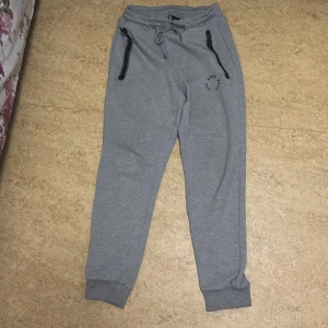 Grå mjukisbyxor från BOSS - Snygga grå mjukisbyxor från BOSS med svarta dragkedjor vid fickorna och snörning i midjan. Byxorna har ribbade muddar vid bensluten och en diskret BOSS-logga tryckt på ena benet. Perfekta för chill eller träning.