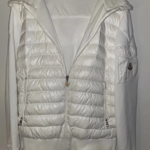 Vit cardigan från Moncler - Säljer en snygg vit cardigan från Moncler med quiltad framsida, huva och dragkedja. Jackan har Moncler-logga på ärmen och praktiska fickor med dragkedja.