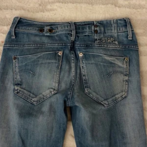 Blå bootcut jeans från G-Star - Säljer ett par snygga blå jeans från G-Star med bootcut passform. Jeansen har slitna detaljer, broderad logga vid bakfickan och klassisk femficksdesign. De är tillverkade i jeansmaterial och har dubbla knappar i midjan för extra stil.