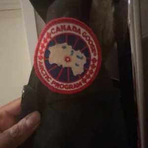 Svart Canada Goose parkas med päls - Svart jacka från Canada Goose med ikonisk röd och vit patch på ärmen. Jackan har luva med beige päls, dragkedja och knappar framtill samt stora fickor. Perfekt för kalla dagar med sin långa passform och snygga detaljer. Köpt på plick💕