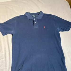 Mörkblå pikétröja från Ralph Lauren - Klassisk mörkblå pikétröja från Ralph Lauren med röd broderad logga på bröstet. Tröjan har korta ärmar, krage och knappar framtill. Tillverkad i mjuk bomull som är skön mot huden.