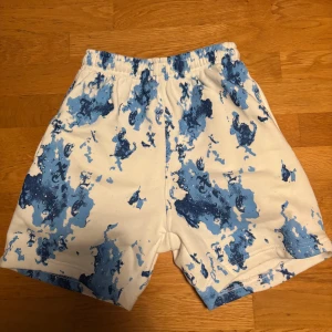 Blåvita shorts från Excite XS - Säljer ett par feta shorts från Excite i storlek XS. De är vita med blått splash-mönster och har resår i midjan. Materialet är mjukt och skönt, perfekt för sommaren. Snygg och avslappnad passform, passar dig som gillar streetwear vibes. Dessa går inte att få tag på längre då det var en begränsad upplaga på just dessa! 