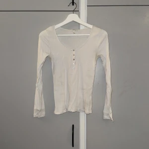 Vit ribbad långärmad topp med knappar - Säljer en vit långärmad topp från H&M med ribbad struktur och rundad hals. Toppen har fem silvriga knappar framtill och är figurnära i modellen. Perfekt att styla med jeans eller kjol för en clean look.