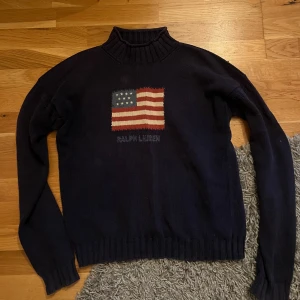 Mörkblå polotröja Ralph Lauren - Snygg mörkblå polotröja från Ralph Lauren med stickad amerikansk flagga på bröstet och broderad logga under. Tröjan har ribbad polokrage, muddar och är tillverkad i mjukt stickat material. Perfekt för dig som gillar klassisk stil med en twist.