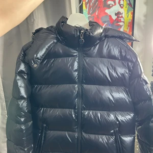 Svart Moncler pufferjacka med dragkedja - Svart pufferjacka från Moncler med glansig finish och klassisk logotyp på ärmen. Jackan har hög krage, två sidofickor med dragkedja och coolt serietidningsfoder på insidan. Perfekt för dig som vill ha en stilren och varm jacka med exklusiv känsla.