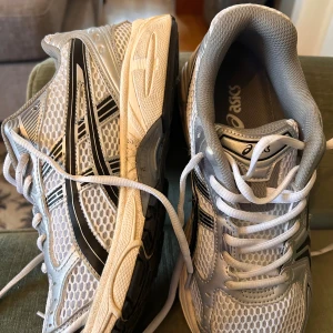 Silvriga Asics sneakers med mesh - Säljer ett par snygga Asics sneakers i silver och vitt med svarta detaljer. Skorna har en sportig design med ventilerande mesh och slitstark syntet på ovandelen. Yttersulan är svart och vit med bra grepp, och de har klassisk snörning för perfekt passform.