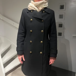 Svart kappa från Ralph Lauren - Svart kappa från Ralph Lauren med dubbelknäppning och snygga guldfärgade knappar med RL-logo. Klassisk krage, figurnära passform och dekorativ knappdetalj bak. Materialet är ull, nylon och kashmir med polyesterfoder. Perfekt för dig som gillar stilrena och tidlösa plagg. Stl 6 motsvarar stl 38. Nypris 5795kr