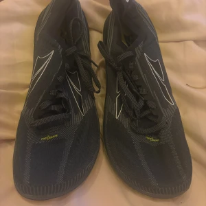 Svarta Altra sneakers strl 42.5 - Säljer ett par svarta Altra sneakers med coola vita detaljer och gul insida. Skorna har snörning och en bred, platt sula med gul och brun mönstrad undersida. Ovandelen är i syntetmaterial med stickad känsla och rund tå. Perfekta för dig som gillar att träna eller bara vill ha bekväma skor.