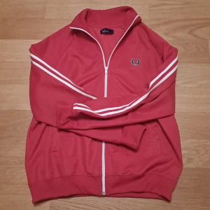 Fred Perry ziptröja - Säljer en röd ziptröja från Fred Perry med vita ränder längs ärmarna och klassisk logga på bröstet. Tröjan har hel dragkedja och sportig look. Perfekt för dig som gillar retrostil och streetwear.