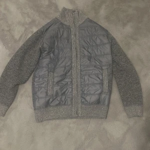 Grå quiltad cardigan jacka - Snygg grå cardigan jacka med quiltad framsida i mörkgrått och stickade ärmar och krage. Insidan har ett coolt svartvitt camouflage-mönster i fleece. Jackan har dragkedja och ribbade muddar vid ärmslut och nederkant. Perfekt för dig som gillar streetstyle och vill ha något unikt.