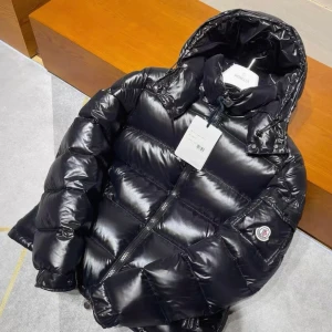 Svart pufferjacka från Moncler - Svart, glansig pufferjacka från Moncler med huva och dragkedja framtill. Jackan har Moncler-logga på ärmen och quiltad design. Insidan är grå och jackan har snörning vid huvan för extra stilpoäng. Perfekt för kalla dagar när du vill se cool ut.