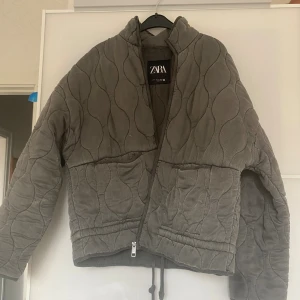 Quiltad grå jacka från Zara - Quiltad jacka från Zara i grått med snyggt vågigt mönster. Jackan har dragkedja, två stora fickor framtill och en hög krage. Materialet är mjuk manchester som ger en chill vibe. Perfekt för dig som gillar loose fit och vill ha något som sticker ut.