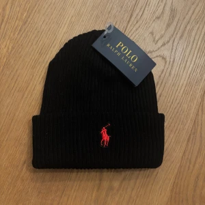 Svart ribbad mössa Polo Ralph Lauren - Svart ribbstickad mössa från Polo Ralph Lauren med klassisk röd broderad logga framtill. Tjockt uppvik och stilren design som passar perfekt till kalla dagar. Tillverkad i mjukt material för extra komfort.