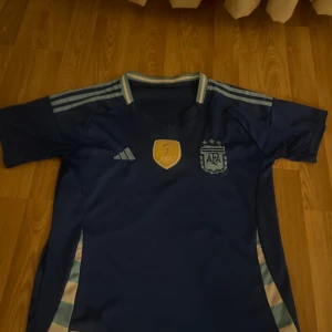 Argentina VM 2022 blå fotbollströja.  - Säljer en officiell Argentina fotbollströja från VM 2022 med FIFA World Champions-märke på bröstet och AFA-emblem. Tröjan är marinblå med ljusblå detaljer och klassiska Adidas-ränder på axlarna. Tillverkad i lätt och ventilerande polyester, perfekt för sommaren.