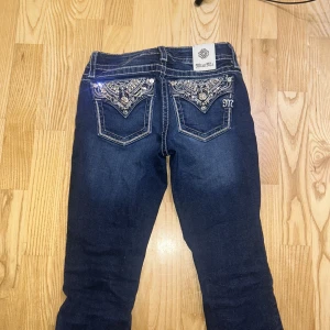 Miss Me jeans med bling bakfickor - Snygga mörkblå jeans från Miss Me med bootcut passform. Bakfickorna är dekorerade med glittriga stenar, broderier och metallknappar. Jeansen har kontrastsömmar och ett litet M broderat på fickan. Materialet är bomull, polyester och elastan för skön stretch. Sitter bra på mig som brukar ha storlek 32