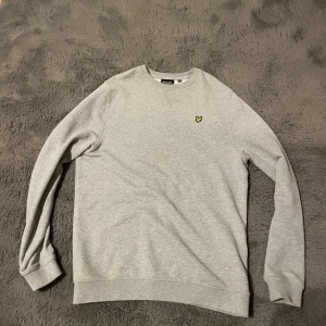 Grå sweatshirt från Lyle & Scott - Snygg ljusgrå sweatshirt från Lyle & Scott med klassisk rund hals och gul logga på bröstet. Tröjan har långa ärmar och ribbade muddar vid ärmslut och nederkant. Perfekt för en chill och stilren look. Den är knappast använd. Säljer den då den är för stor!
