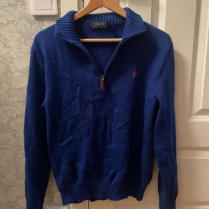 Blå stickad tröja Polo Ralph Lauren - Blå stickad tröja från Polo Ralph Lauren med half zip och ribbad krage. Klassisk röd logga broderad på bröstet. Långärmad modell med ribbade muddar och nederkant. Perfekt för dig som gillar stilren och tidlös design.