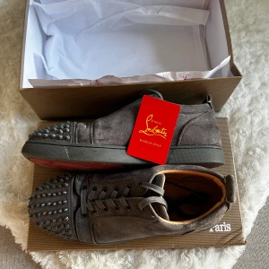 Grå Christian Louboutin sneakers med nitar - Säljer ett par gråa sneakers från Christian Louboutin i mocka med coola nitar på tån och klassisk röd sula. Skorna har snörning och en rund tå, vilket ger dem en edgy vibe. Kommer med originalkartong och tagg. Perfekt för dig som gillar statement pieces.