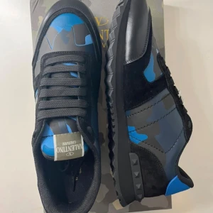 Valentino Rockrunner sneakers blå/svart - Snygga Valentino Rockrunner sneakers med blått och svart kamouflagemönster. Skorna har svarta snören, detaljer i mocka och skinn samt en cool platt sula med nitar på hälen. Perfekta för dig som vill sticka ut med stil.