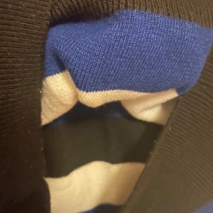 Randig ärmlös stickad tröja V.R.M - Säljer en ärmlös stickad tröja från V.R.M med breda svarta, blå och vita ränder. Tröjan har v-ringning och är perfekt för lager-på-lager-stil. Materialet är mjukt och stretchigt, vilket gör den skön att bära.