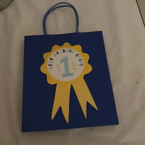 Blå presentpåse med studenttema - En blå presentpåse med handtag och en stor gul rosett framtill med texten 'Best Student Ever!' och en etta i mitten. Perfekt för att fira någon som tagit studenten eller gjort något extra bra i skolan.