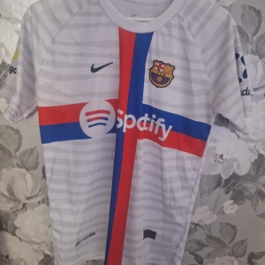 Barcelona Pedri 16 vit matchtröja Nike - Säljer en FC Barcelona fotbollströja med Pedri 16 på ryggen. Tröjan är vit med tunna grå ränder, har röda och blå detaljer samt klubbmärke och Nike-logga på bröstet. Dri-FIT material som andas, kortärmad och med Spotify som sponsortryck.