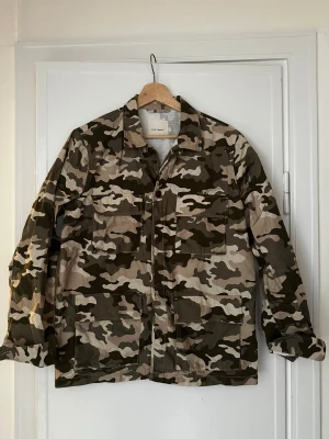 Camo overshirt från A Day's March - Snygg overshirt i camomönster från A Day's March. Jackan har fyra stora fickor framtill, klassisk krage och knäppning. Tillverkad i ett slitstarkt bomullstyg, perfekt för lager-på-lager. Färgerna går i grönt, beige och brunt.
