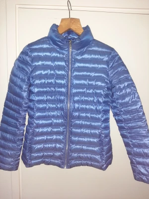 Blå pufferjacka från Massimo Dutti - Snygg blå pufferjacka från Massimo Dutti med glansig finish och horisontella sömmar. Jackan har hög krage och dragkedja framtill. Perfekt för kalla dagar och enkel att matcha med olika outfits.