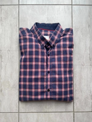 Mörkblå-röd rutig skjorta från Mouli’1 - Snygg mörkblå skjorta från Mouli’1 med rött och vitt rutmönster. Skjortan har klassisk krage, knäppning framtill och långa ärmar med manschett. Perfekt för en stilren och avslappnad look. Nypris -:2200. Vid frågor eller funderingar skriv gärna! Strl: XxL, passar L