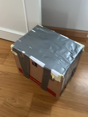 Stor kartong med silvertejp - En rejäl kartong som är förstärkt med flera lager silvertejp runt om. Perfekt för förvaring eller flytt. Kartongen är brun med röda detaljer och har en fyrkantig form.