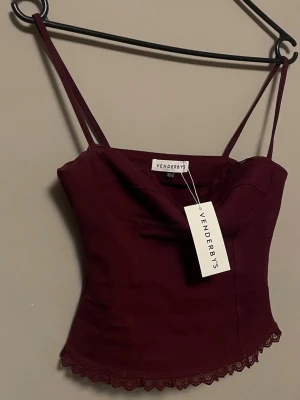 Vinröd korsettopp med spetsdetaljer - Vill man ha fler bilder så skriv! Snygg vinröd korsettopp från venderbys. Toppen har figurnära passform och markerade sömmar som ger en trendig siluett. Perfekt att styla med både jeans och kjol för en cool look. Helt ny. 
