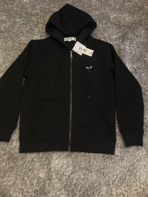 Svart Comme des Garçons Play zip hoodie - Svart hoodie med dragkedja från Comme des Garçons Play. Klassisk design med huva, fickor framtill och det ikoniska hjärtloggan med ögon på bröstet. Perfekt för en stilren och avslappnad look skriv vid eventuella frågor.