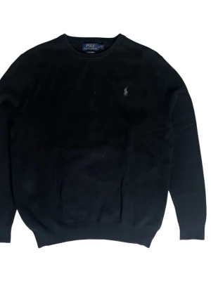 Svart stickad tröja från Polo Ralph Lauren - Klassisk svart stickad tröja från Polo Ralph Lauren med rund halsringning och diskret broderad logga på bröstet. Tröjan har ribbade muddar vid ärmslut och nederkant. Perfekt för en stilren look.