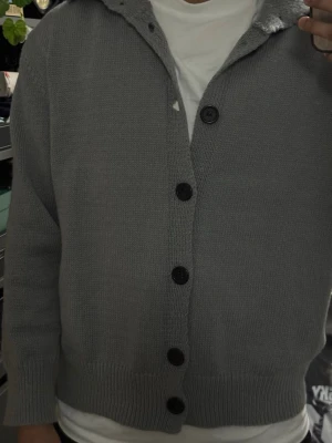 Grå stickad kofta med knappar - Klassisk grå stickad kofta i storlek S med stora svarta knappar framtill. Koftan har ribbade muddar vid ärmslut och nederkant samt en rak passform. Perfekt att slänga över en t-shirt för en chill look. Osäker på märke. Normal passform. 
