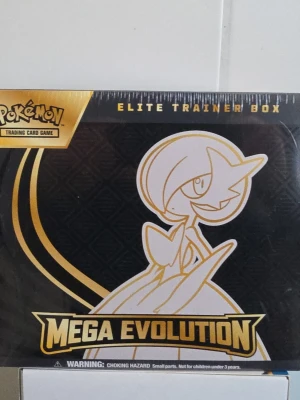 Pokémon Mega Evolution Elite Trainer Box - Säljer en Pokémon Mega Evolution Elite Trainer Box med motiv av Gardevoir i vitt och guld på svart bakgrund. Boxen innehåller booster packs, energi-kort, sleeves och tillbehör för Pokémon Trading Card Game. Perfekt för samlare och spelare som vill ta sitt kortspel till nästa nivå.