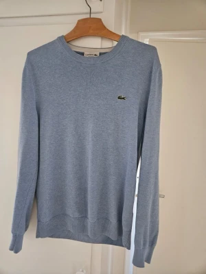 Ljusblå tröja från Lacoste - Snygg ljusblå tröja från Lacoste med klassisk rund halsringning och den ikoniska krokodilloggan på bröstet. Tröjan har långa ärmar och ribbade muddar vid ärmslut och nederkant. Passar inte mig ej andvänd