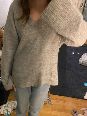 Grå stickad V-ringad tröja - Mysig grå stickad tröja med V-ringning och breda ribbade muddar vid ärmslut och nederkant. Tröjan har en avslappnad passform och är perfekt för lager på lager 🩶lite nopprig 