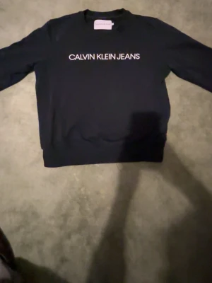 Svart Calvin Klein Jeans sweatshirt - Svart sweatshirt från Calvin Klein Jeans med vit logotyptext på bröstet. Klassisk rund halsringning och långa ärmar. Perfekt för en stilren och avslappnad look.