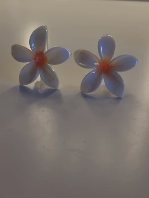 Blommiga hårklämmor i plast - Två söta hårklämmor formade som vita blommor med fem kronblad och en mjukt orange mitt. Klämmorna är gjorda i glansig plast och har en genomskinlig klämma på baksidan. Perfekt för att piffa upp frisyren med en tropisk vibe.